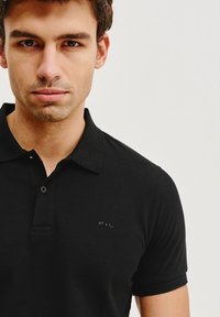 Czarna koszulka polo z bawełny, z klasycznym kołnierzykiem, trzema guzikami i subtelnym logo 'P.L' na lewej stronie klatki piersiowej.