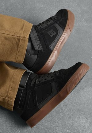 Baskets montantes DC noires avec semelles brunes portées avec un pantalon beige sur un sol gris texturé.