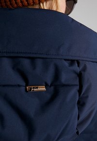 Tmavě modrá prošívaná bunda s kovovou značkovou cedulkou na zádech, zdůrazňující prošívání a hladkou texturu.