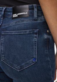 Jeans din denim de un albastru închis cu un aspect uzat. Au o etichetă neagră din piele la talie și un mic 'K' brodat de culoare albastră pe buzunar.