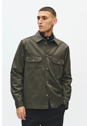 Junger Mann, der nach links schaut, trägt eine dunkelgrüne Hemdjacke mit Knöpfen und zwei Brusttaschen, schwarze Hose und darunter einen dunklen Reißverschluss-Pullover.