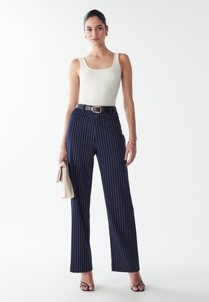 Jūras spēku zilos svītrainos platajās biksēs ar augstu vidukli un melnu jostu. Pāris ar pieguļošu krēmkrāsas ribotu ķermeņa augšdaļu un turot rokā beigu krāsas somu.