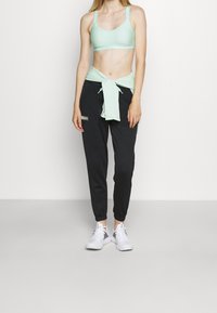 Sujetador deportivo verde claro con una banda con logo, combinado con joggers negros que tienen un parche lateral. Zapatillas deportivas blancas completan el look.