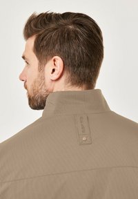 Beige, textured jacket with a stand-up collar. Features a vertical stripe pattern and a tag detail at the back with the brand name.