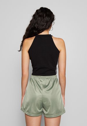 Kobieta o długich ciemnych włosach, ubrana w czarny bezrękawnik i jasne zielone szorty z elastycznym pasem, stojąca tyłem na jednolitym tle.