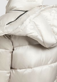 Chaqueta acolchada en beige claro, con una textura brillante, secciones acolchadas y un bolsillo lateral con cremallera y herrajes negros.