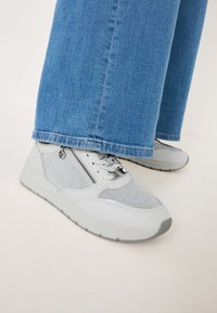 Graue Sportschuhe aus Mesh und synthetischen Materialien, mit einem seitlichen Reißverschluss, strukturierten Akzenten und einer Gummisohle. Getragen mit blauen Jeans.