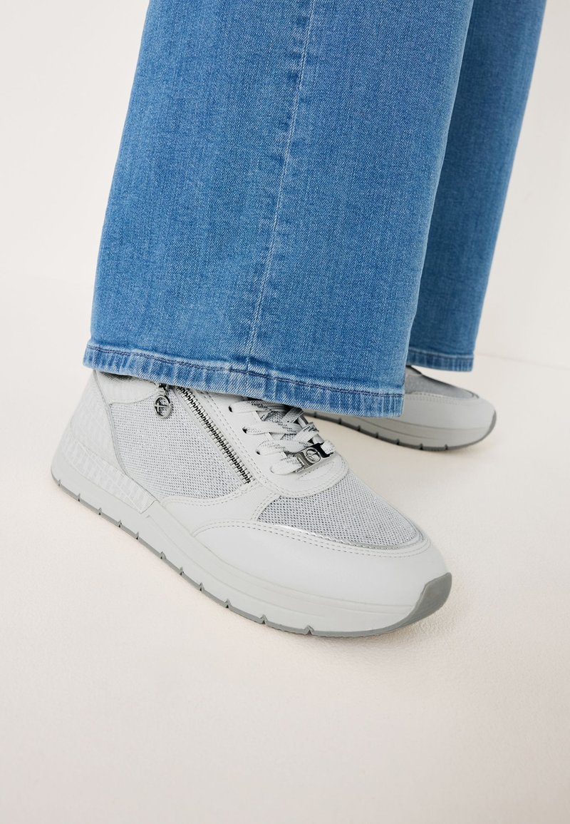 Graue Sportschuhe aus Mesh und synthetischen Materialien, mit einem seitlichen Reißverschluss, strukturierten Akzenten und einer Gummisohle. Getragen mit blauen Jeans.