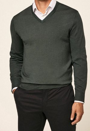 Mann trägt dunkelgrünen V-Ausschnitt-Pullover über weißem Hemd mit schwarzer Hose, posiert mit einer Hand in der Hosentasche vor einfarbigem Hintergrund.