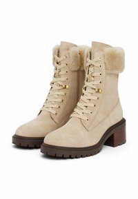 Botines de ante beige con un cuello de pelo sintético, cordones en la parte delantera, acentos de hardware dorado y un tacón grueso marrón. Suela de goma texturizada.