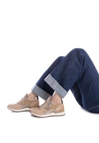 Zapatillas de ante beige con acentos metálicos, punta redonda y cremallera lateral. Llevadas con jeans de mezclilla azul oscuro, con puños enrollados y dobladillos en contraste.