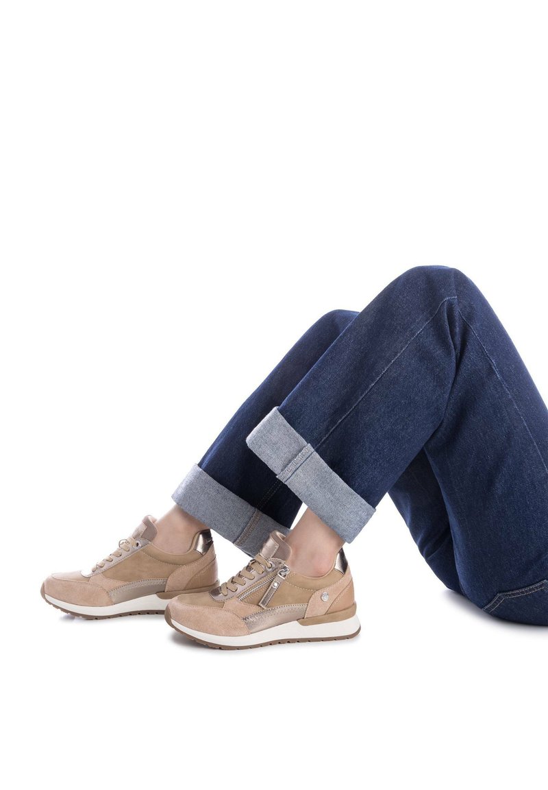 Zapatillas de ante beige con acentos metálicos, punta redonda y cremallera lateral. Llevadas con jeans de mezclilla azul oscuro, con puños enrollados y dobladillos en contraste.