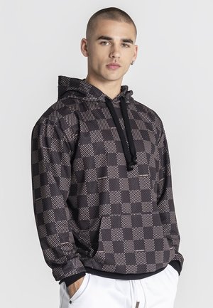 Mladý muž stojaci s rukami v vreckách s čierno-šedou károvanou mikinou s kapucňou so šnúrkami a bielymi nohavicami na jednoduchom pozadí.