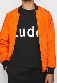 Oranssi pommitakki vetoketjulla, resorikauluksella ja -hihoilla, päällystetty mustan t-paidan päälle, jossa on suuret valkoiset tekstit. Sileä pinta.