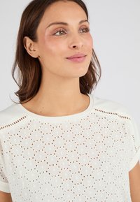 Camiseta de manga corta de cuello redondo en un suave color crema, con un patrón de ojales abiertos y tejido texturizado. Presenta costuras de acento a lo largo del escote.