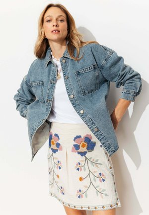 Femme portant une veste en denim bleue par-dessus une chemise blanche et une jupe blanche avec une broderie florale colorée, posant devant un fond blanc.
