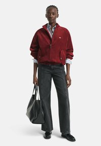 Veste en velours côtelé rouge avec fermeture éclair, associée à une chemise bleu clair, un pantalon en denim noir et un sac fourre-tout noir texturé. Des chaussures décontractées complètent la tenue.