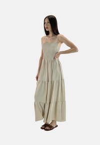 Abito maxi beige a strati con corsetto smock e sottili spalline, realizzato in un tessuto morbido che offre una silhouette comoda e fluida.
