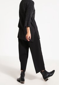 Tunique tricotée longue gris foncé avec une coupe décontractée et des fentes latérales, associée à un pantalon noir à jambes larges et des bottines à talon compensé.
