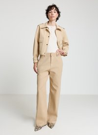 Beige kurz geschnittene Jacke mit zwei Fronttaschen, kombiniert mit weiten beigen Hosen und spitz zulaufenden Absatzschuhen mit Schlangenprint. Glatter, strukturierter Stoff.