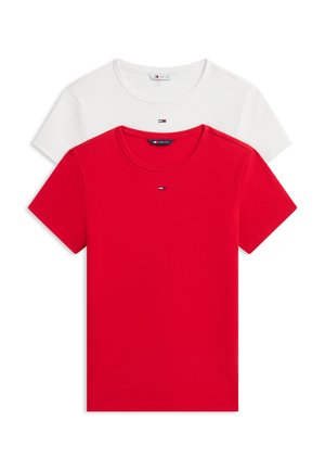 Femme aux cheveux noirs bouclés portant un t-shirt rouge, un pantalon blanc, une ceinture noire et des chaussures foncées, tenant un sac à main marron avec une écharpe à motifs.