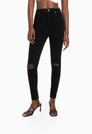 Femme portant un jean skinny noir taille haute avec des genoux déchirés et des sandales à talons hauts noires à lanières, debout devant un fond blanc.