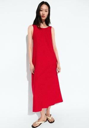 Donna in piedi con un vestito midi rosso senza maniche e sandali beige infradito su uno sfondo bianco uniforme.