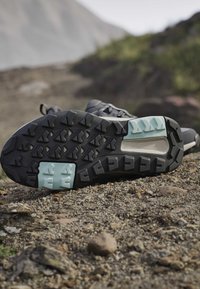 Semelle d'une chaussure de randonnée noire et bleu clair sur un sentier rocheux avec un paysage montagneux en arrière-plan flou.