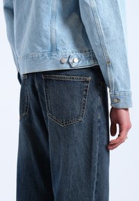 Chaqueta de mezclilla azul claro con botones a presión plateados, llevada sobre jeans de mezclilla azul oscuro con bolsillos traseros y un acabado de tela texturizada.