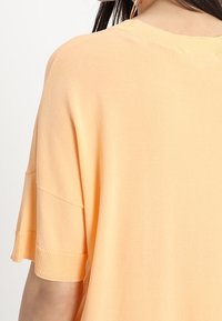 Vrouw in een losse, kortame trui in perzikkleur met ribgebreide mouwen en een ronde halslijn, weergegeven van de achterkant.