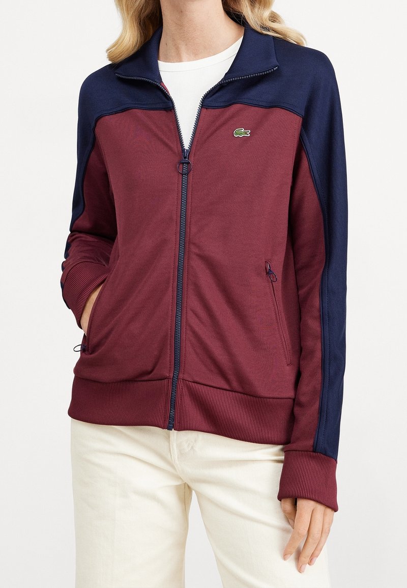 Femme portant une veste zippée bordeaux avec des épaules et manches bleu marine, associée à un pantalon clair, les mains dans les poches.