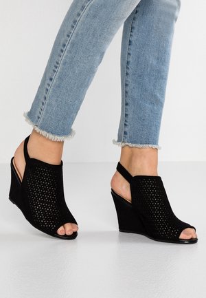 Voeten in lichtblauwe franje jeans met een zwarte open teen wedge hiel met geometrisch uitsnijdontwerp.