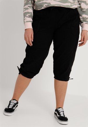 Osoba ubrana w czarne skrócone spodnie z bocznymi kieszeniami i czarno-białe trampki, stojąca na jasnym tle.