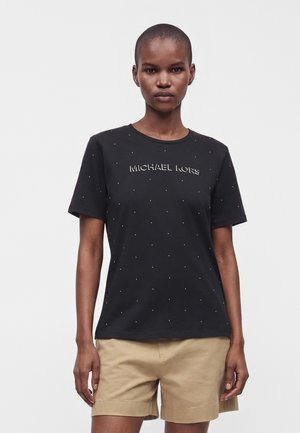 Modelka w czarnej koszulce Michael Kors z małymi kropkami i beżowych szortach, stojąca na jednolitym białym tle.