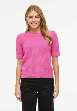 Donna che indossa un maglione a maniche corte di colore rosa acceso e dalla trama evidente, con pantaloni neri, sorridente su uno sfondo bianco neutro.