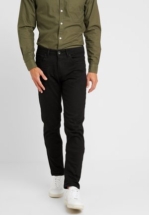 Zwarte slim-fit jeans met een gladde textuur, gecombineerd met een olijfgroene knoopshirt. De outfit wordt aangevuld met witte sneakers.