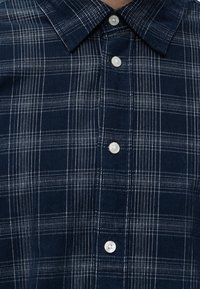 Chemise à carreaux bleu foncé avec des rayures blanches verticales et horizontales, avec un col et quatre boutons blancs sur le devant.
