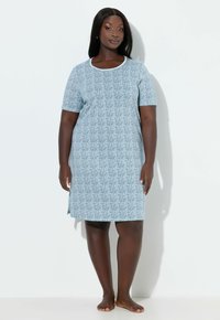 Mujer descalza, de pie, vistiendo un vestido azul claro con estampado, de manga corta y hasta la rodilla, sobre un fondo blanco liso.