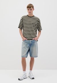Camisa de manga corta a rayas en verde oliva oscuro y blanco, combinada con pantalones cortos de mezclilla azul claro. El modelo lleva zapatillas blancas con detalles en negro.