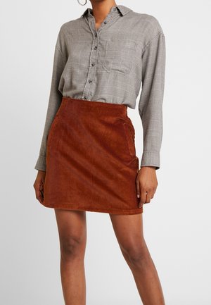 Kobieta w szarej koszuli w kratę zapinanej na guziki, wpuszczonej w rdzawobrązową mini spódnicę z sztruksu z bocznymi kieszeniami, stojąca na jednolitym tle.