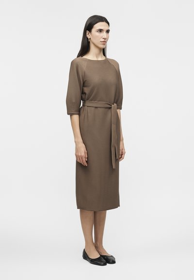 Γυναίκα που στέκεται φορώντας καφέ ριμπ μίντι φόρεμα με μανίκια τριών τετάρτων, ζώνη που ταιριάζει, χρυσούς κρίκους σκουλαρίκια και μαύρα μπαλαρίνες.