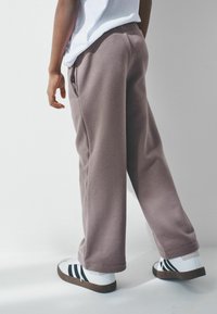 Gai brūni brīvi piegulstoši sporta bikses ar sānu kabatām, elastīgu jostu un plaša kroņa dizainu, apvienoti ar baltiem sporta apaviem ar melnām svītrām.