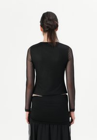 Black long-sleeve top with sheer sleeves, lightweight fabric, and a relaxed fit. Worn over a black textured skirt, viewed from the back.