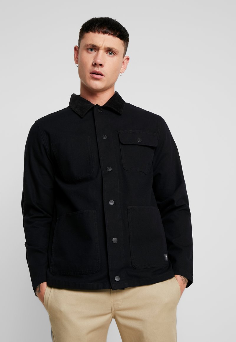 Black jacket with a corduroy collar, front snap buttons, and two chest pockets. Made of a sturdy material with a smooth texture.