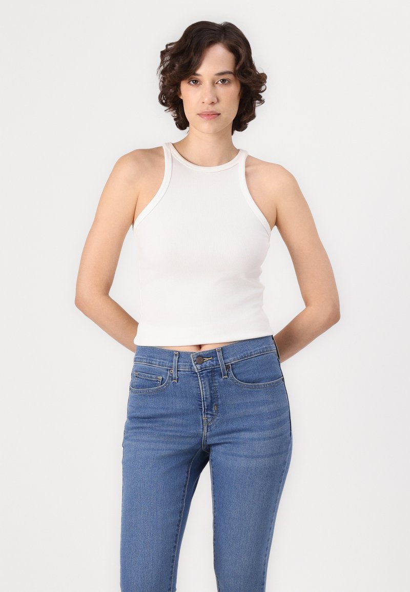 Femme avec des cheveux courts et bouclés portant un haut sans manches blanc et un jean taille haute bleu, se tenant devant un fond blanc uni.