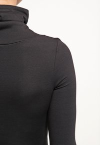 Chemise noire à col roulé à manches longues portée, montrant le dos et l'épaule sur un fond clair uni.