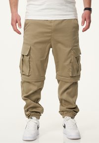 Khakifarbene Cargo-Hosen mit lockerer Passform, mehreren Seitentaschen, elastischen Bündchen und glatter Textur. Getragen mit weißen Sneakern.