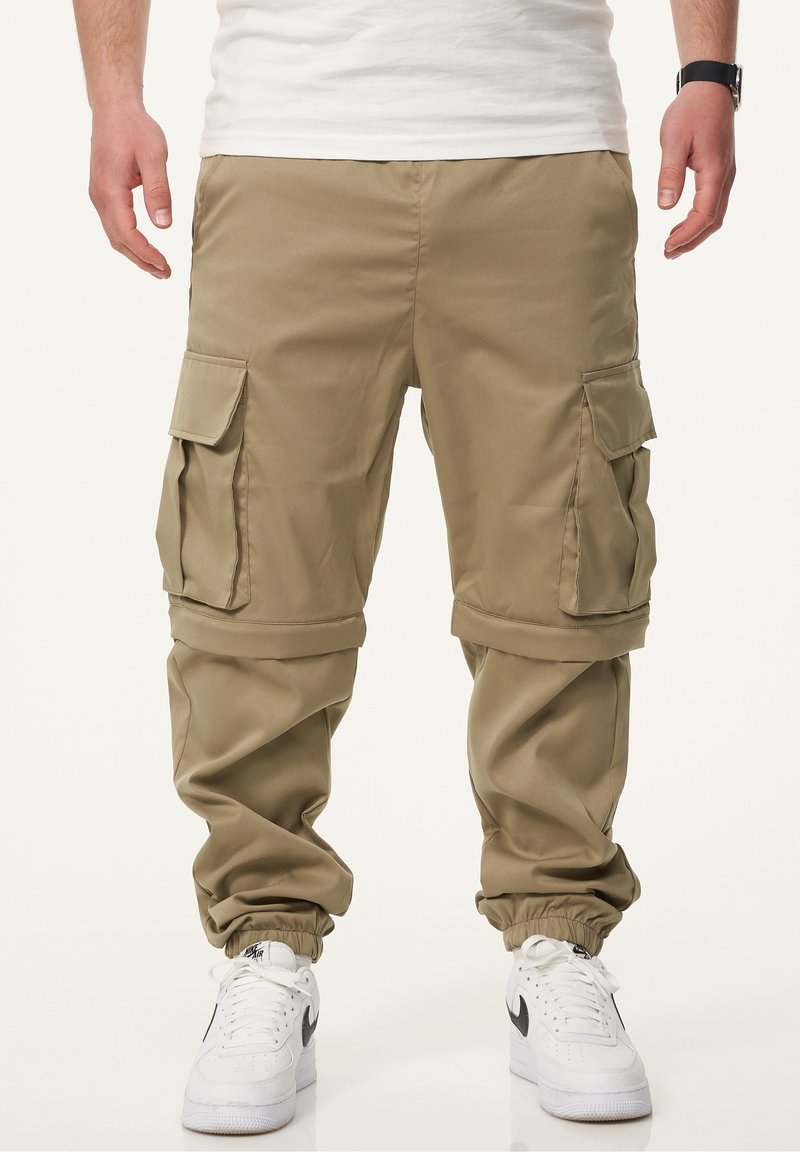 Khakifarbene Cargo-Hosen mit lockerer Passform, mehreren Seitentaschen, elastischen Bündchen und glatter Textur. Getragen mit weißen Sneakern.