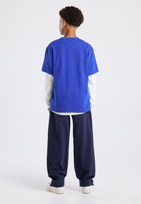 Blue cotton t-shirt layered over a white long-sleeve shirt, paired with loose navy pants and white sneakers, shot from behind.