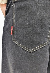 Pantalones de mezclilla negra que muestran un bolsillo trasero con costuras amarillas y una etiqueta roja. La tela tiene una apariencia texturizada.